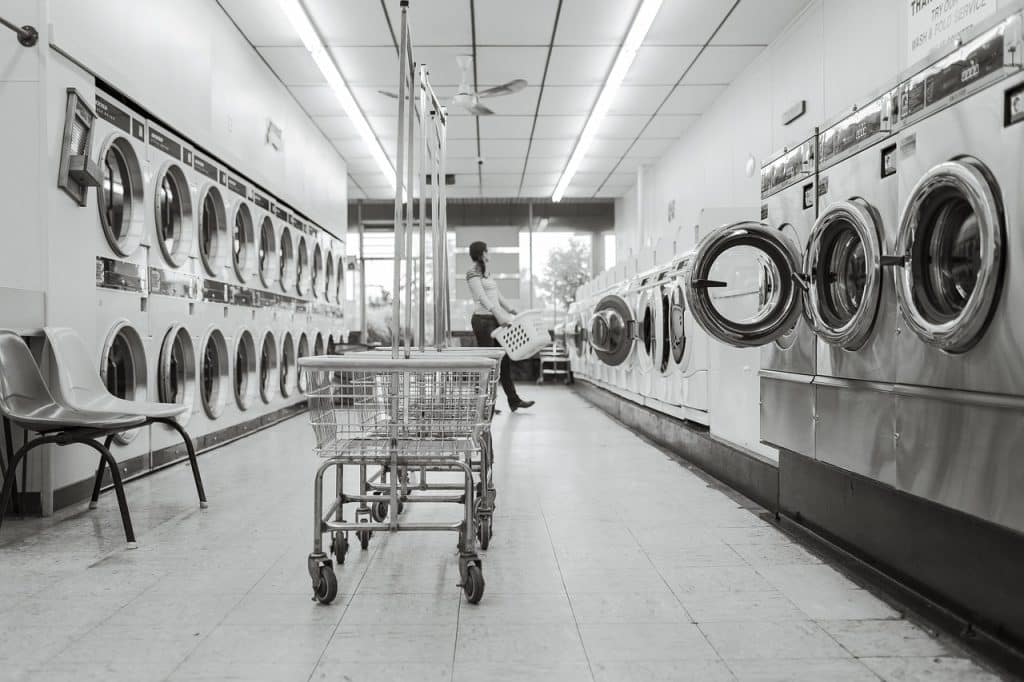 A monochrome image capturing the serene ambiance of a spacious laundromat. Multiple washing machines line the walls while a solitary individual tends to their laundry.